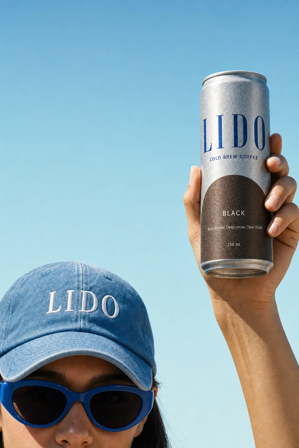 A raised hand holding a LIDO Black can up to the sky beside the blue cap and sunglasses.