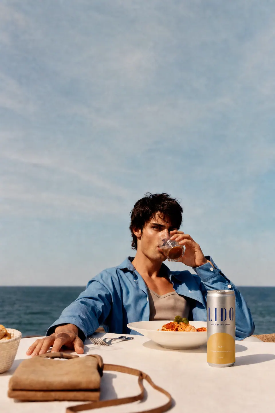 A man in a blue shirt drinking at a seaside lunch table beside a LIDO Oat can and a bowl of pasta.
