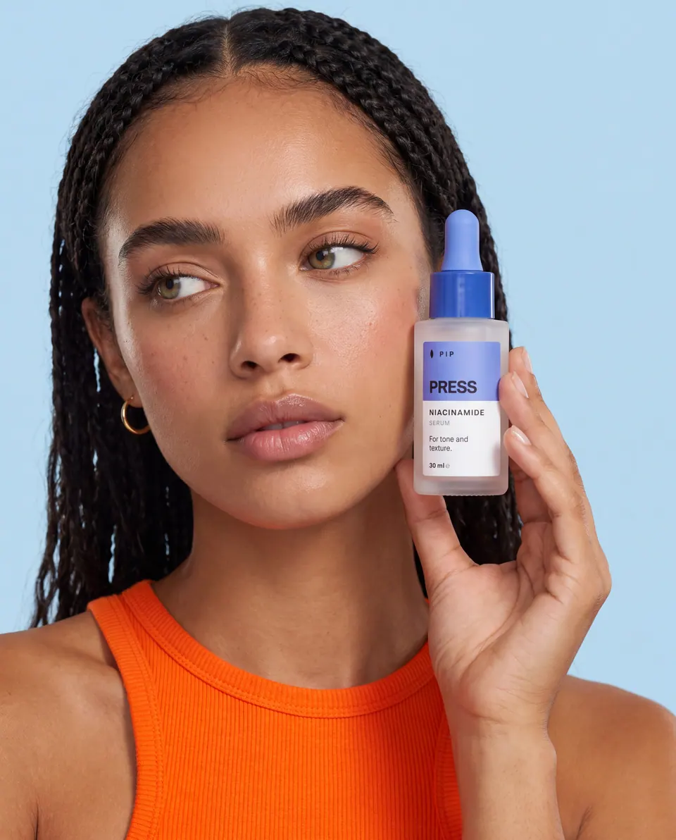 Model with braided hair and an orange ribbed top holding a PRESS bottle beside her cheek against pale blue.